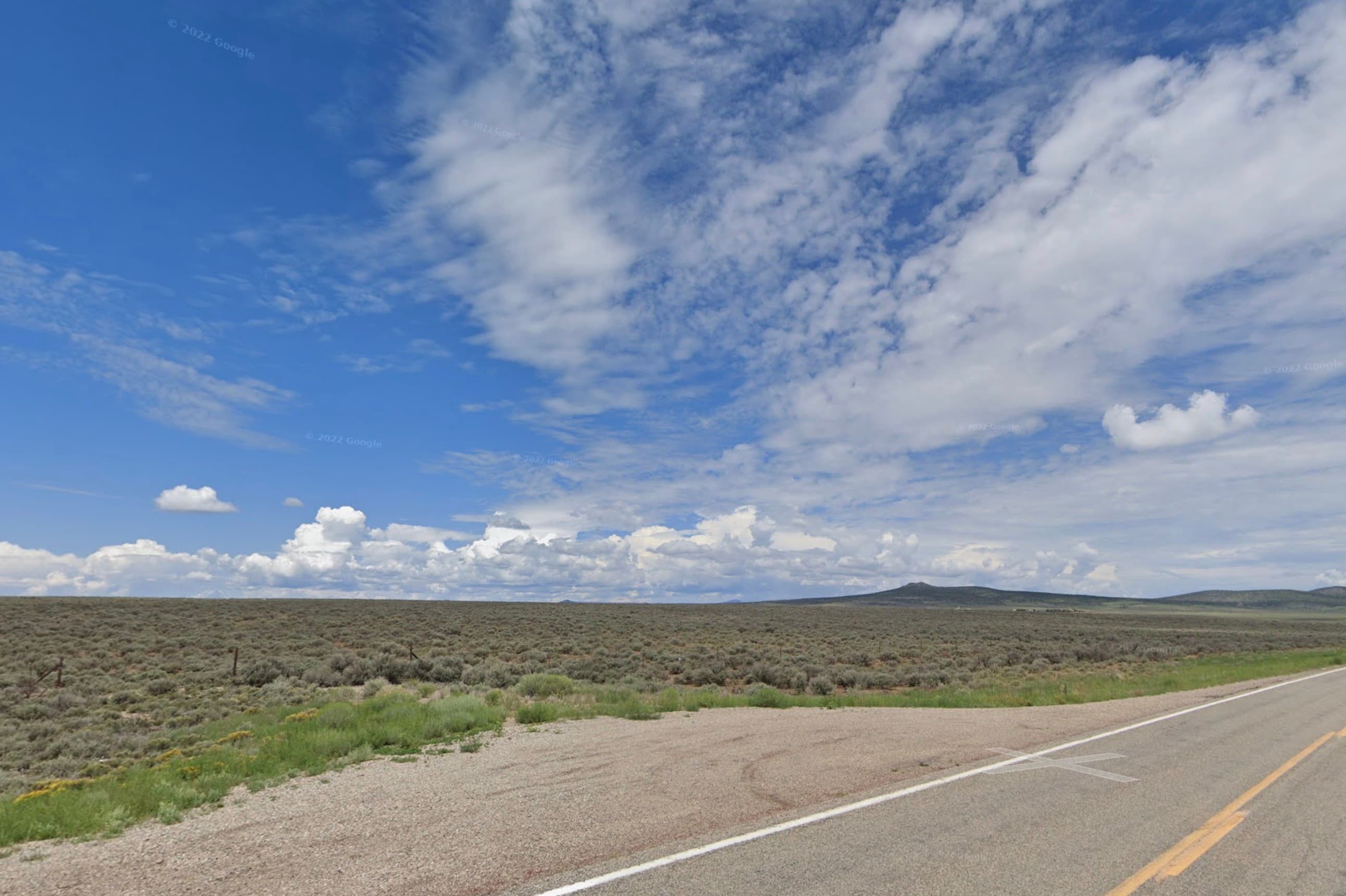 Two-lane highway with sagebrush on both sides and mountains across the valley in Taos County, New Mexico.