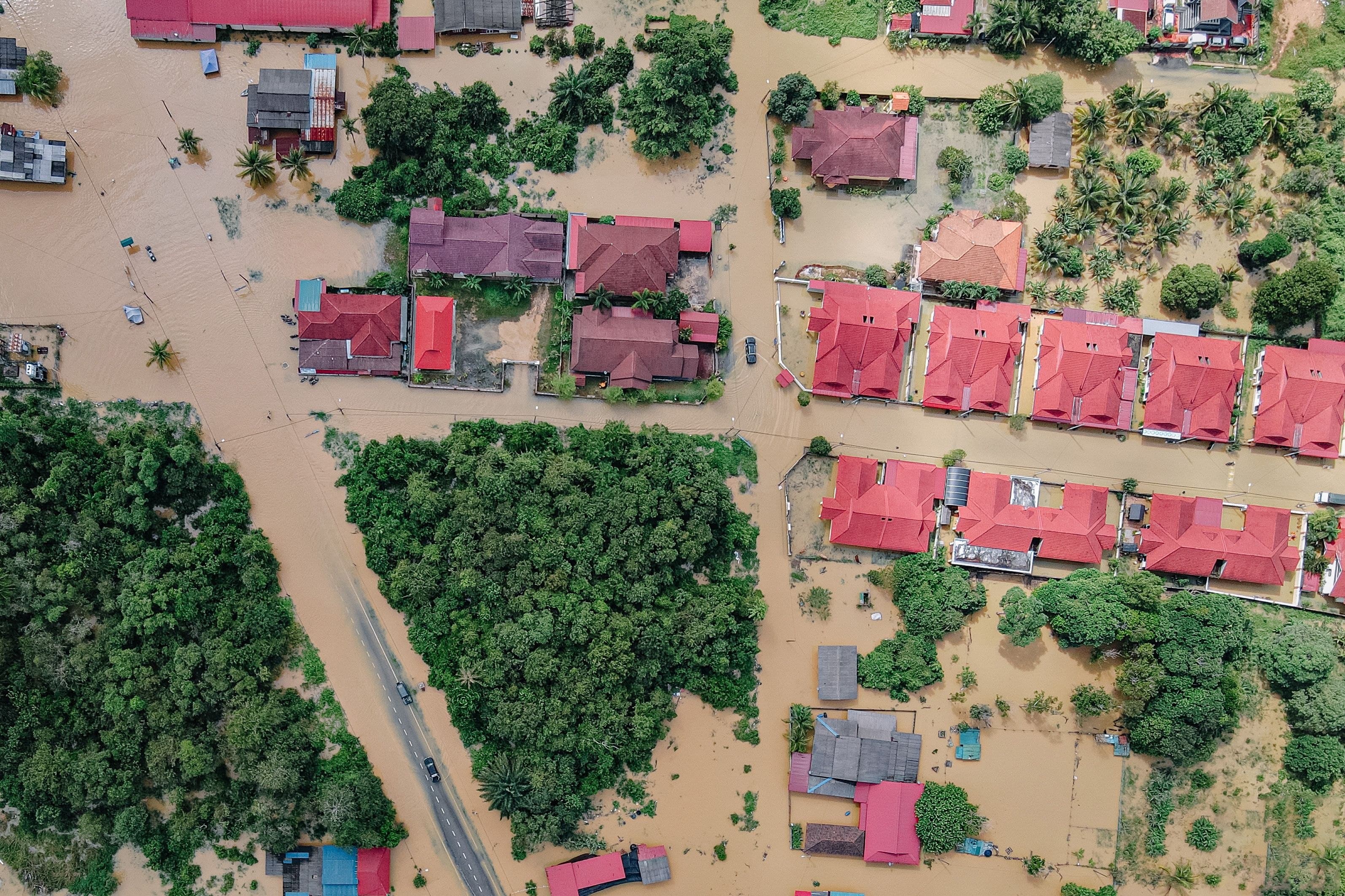 Aerial view of a flooded neighbourhood, brown water standing between rows of red-roofed houses and green trees.