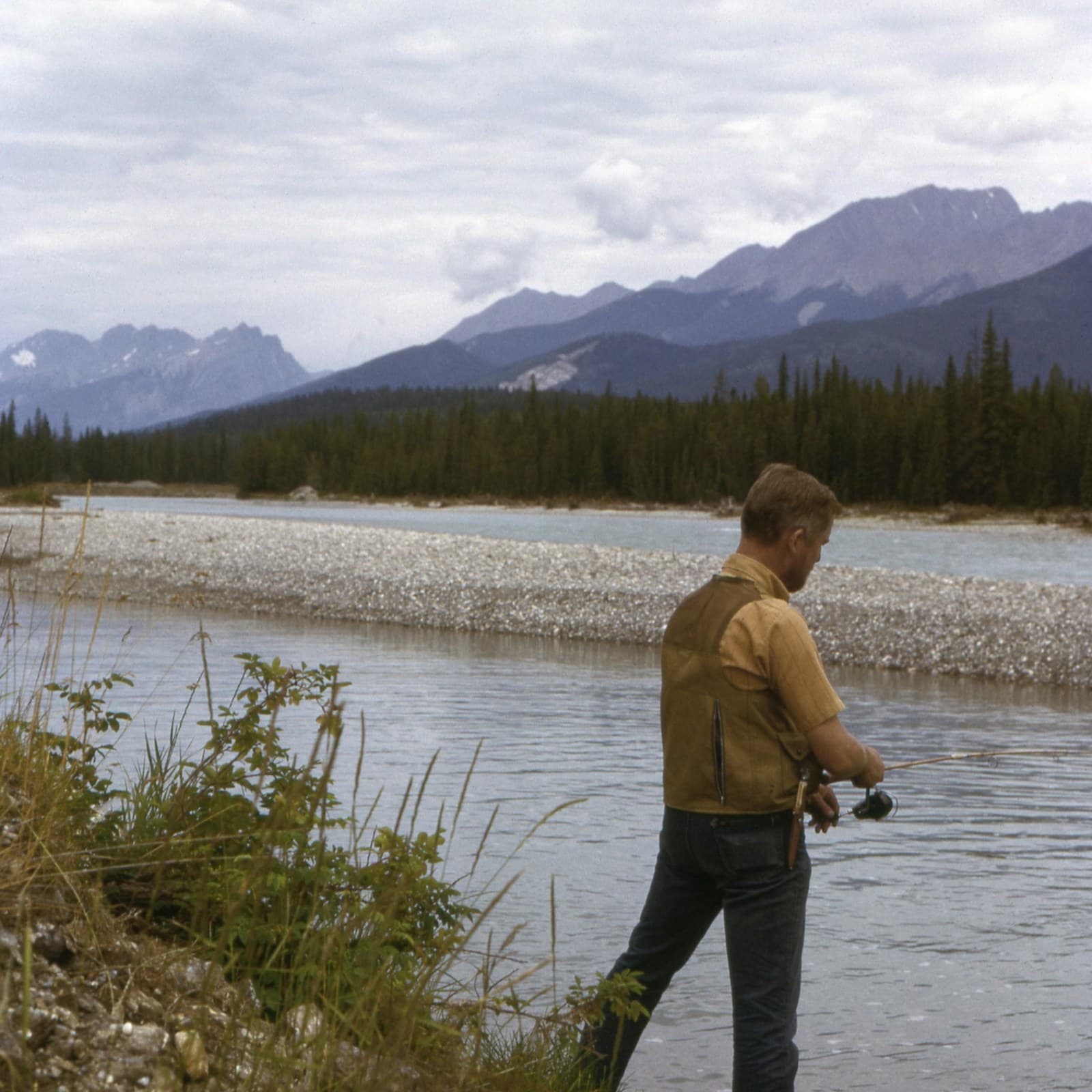 An angler in a canvas jacket working a reel at the edge of a wide river, gravel bar and snowy peaks beyond.