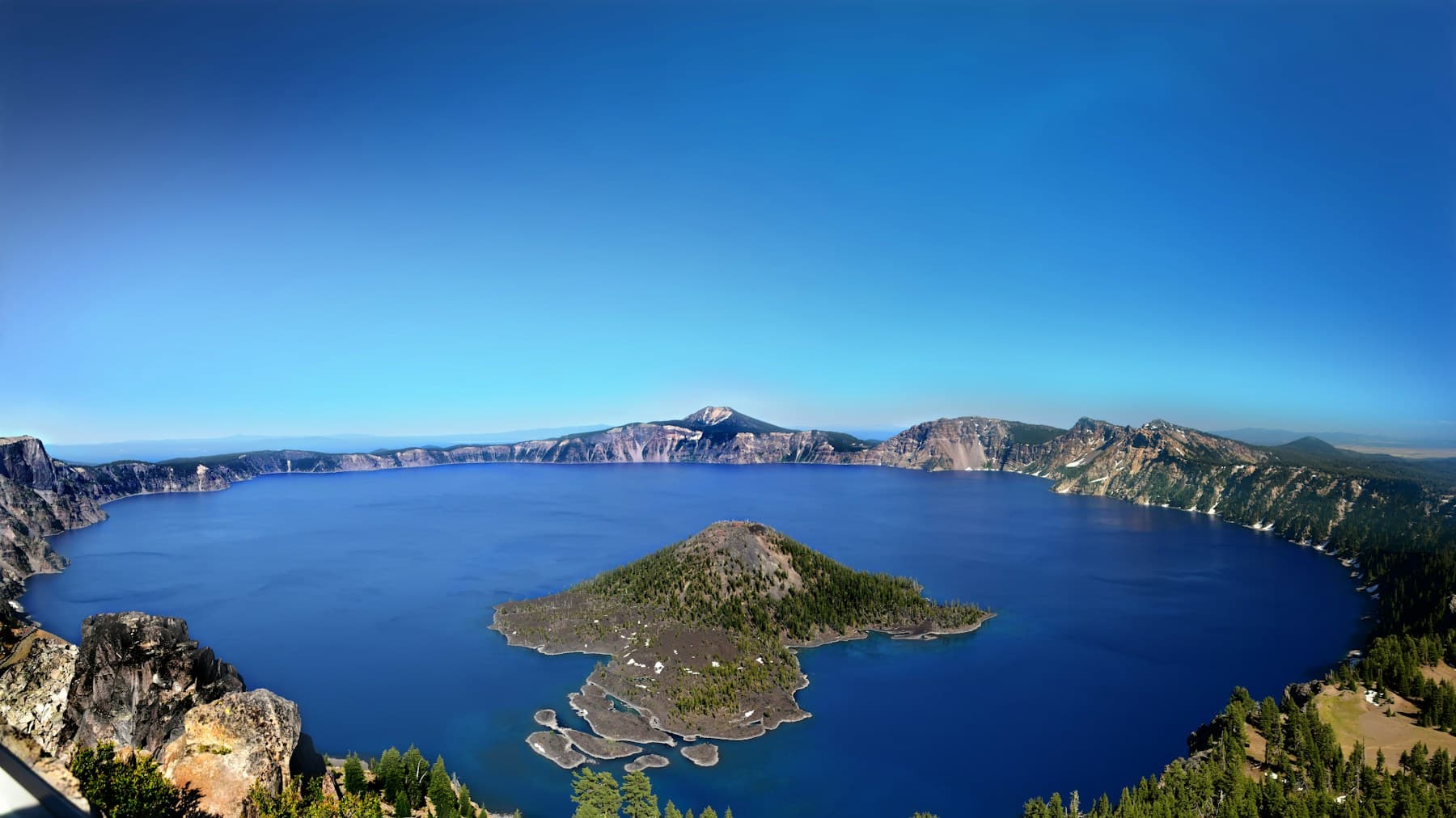 Crater Lake seen from the caldera rim, deep blue water ringed by steep cliffs with Wizard Island rising near the western shore.