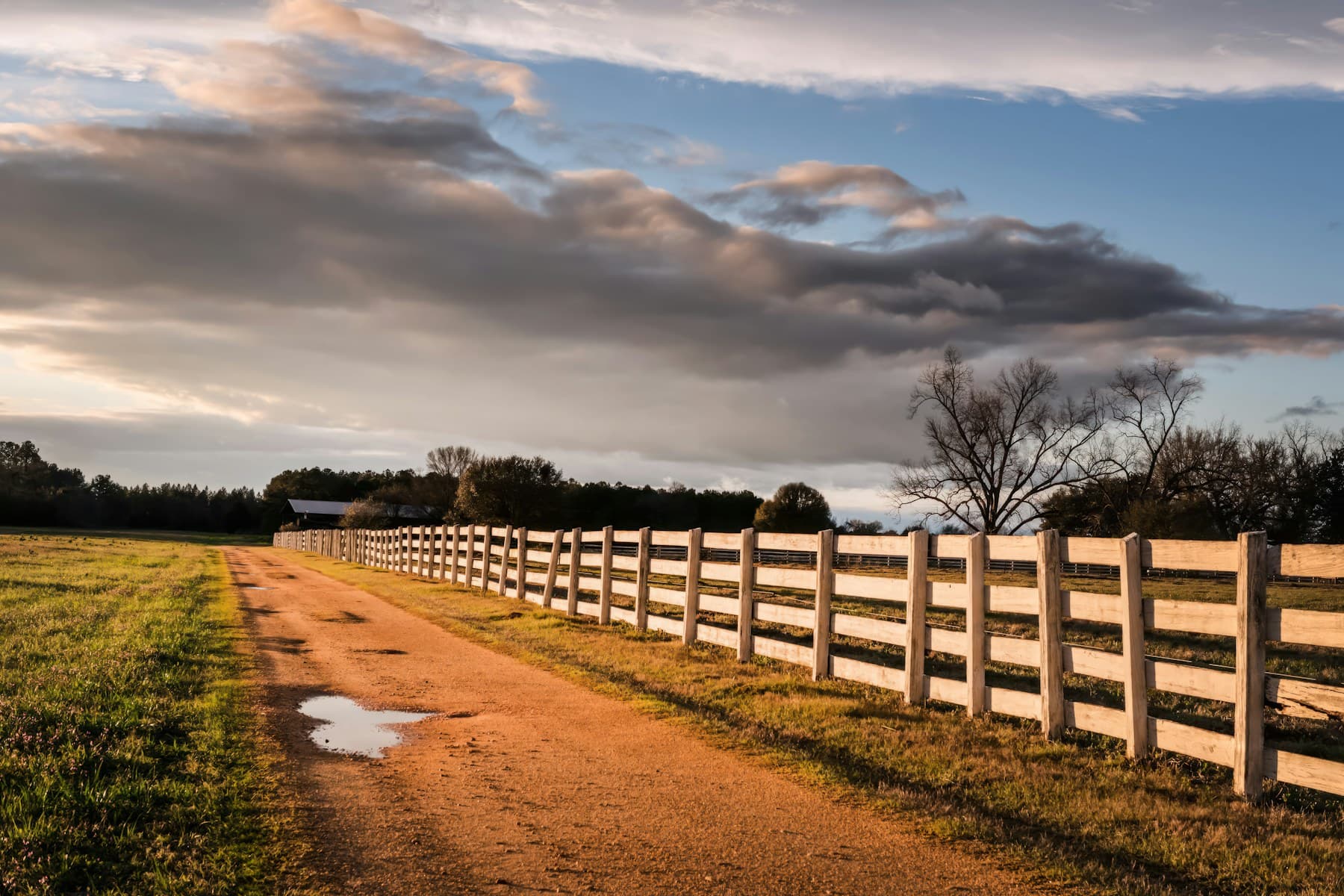 A dirt farm road running beside a long wooden rail fence under a wide bank of evening cloud.