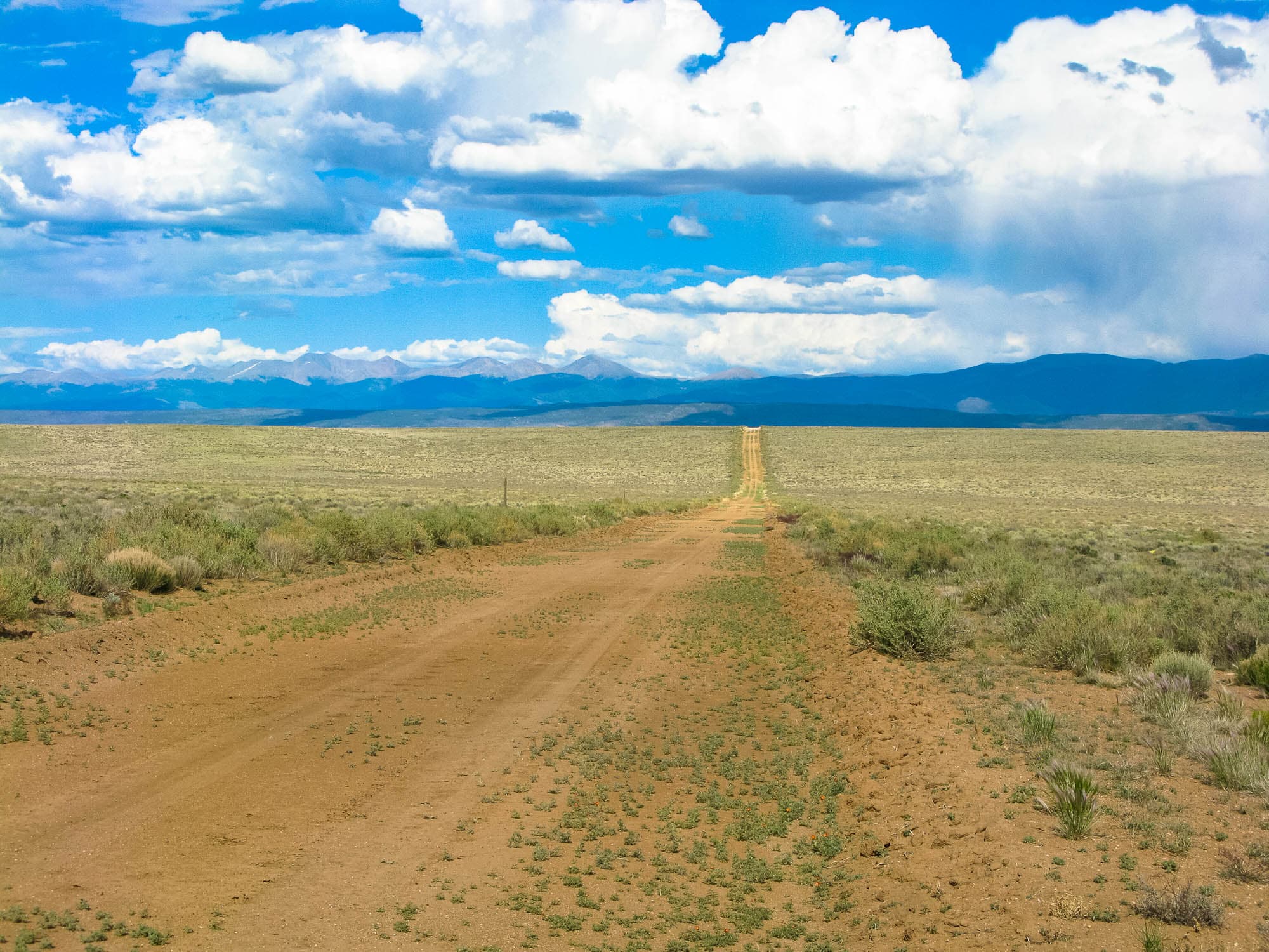 A graded dirt road running dead straight to the horizon across open sage flats, snow-dusted peaks behind and tall cumulus stacked overhead.