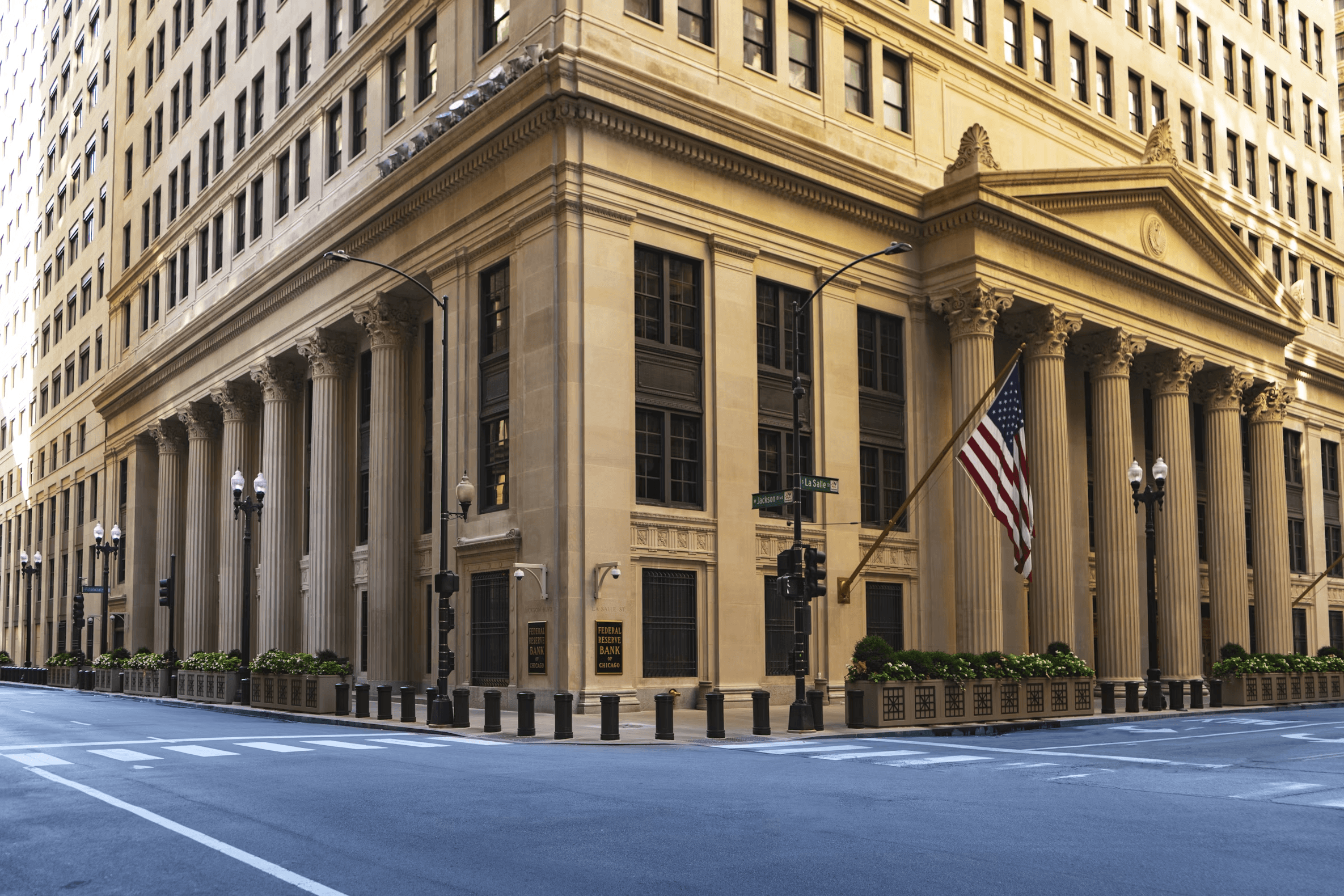 A grand stone bank building with tall columns on a city corner, an American flag hanging above the empty street.