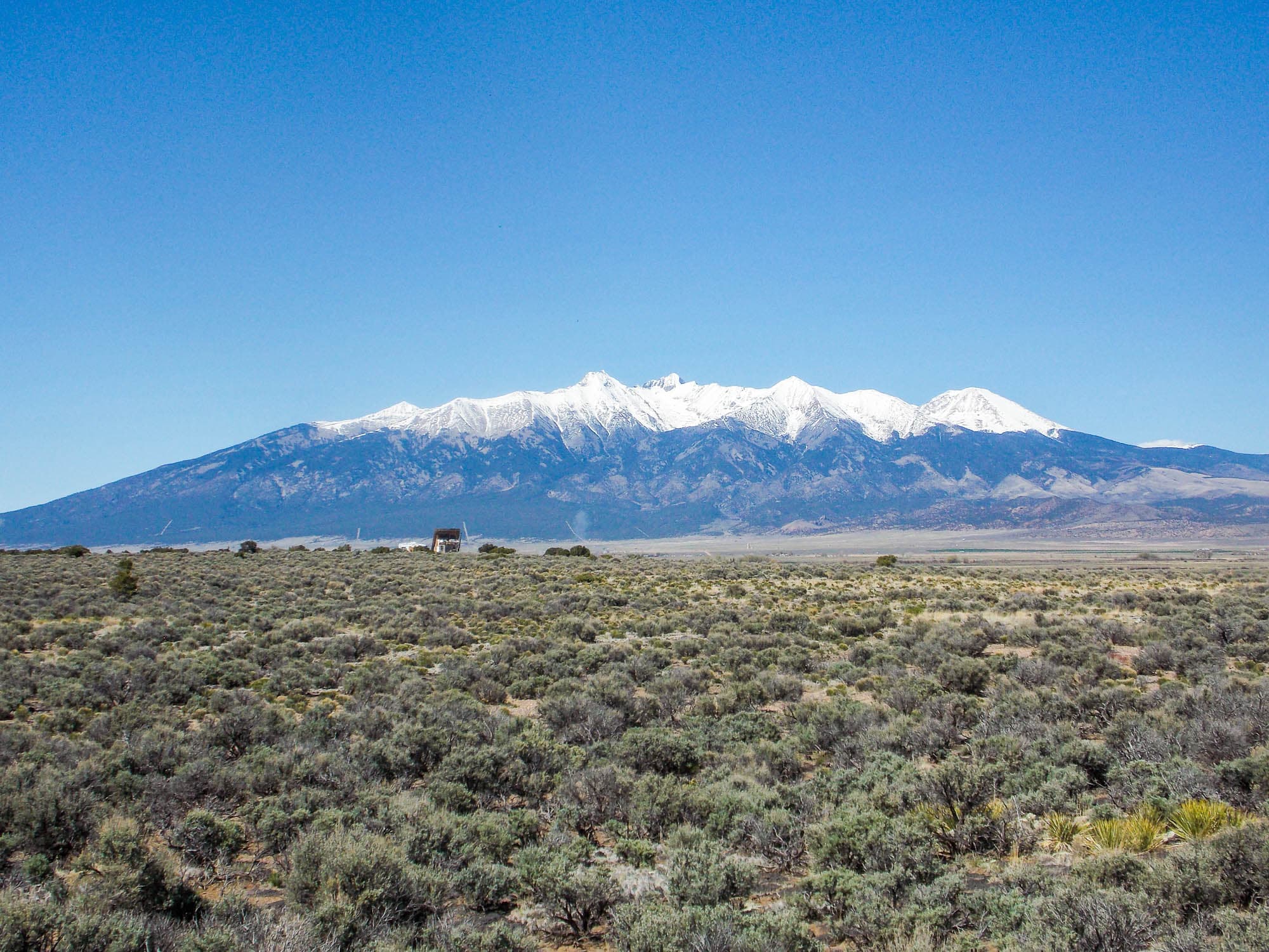 A long snow-capped massif rising above a wide sage flat under deep blue sky, one small building at its base.