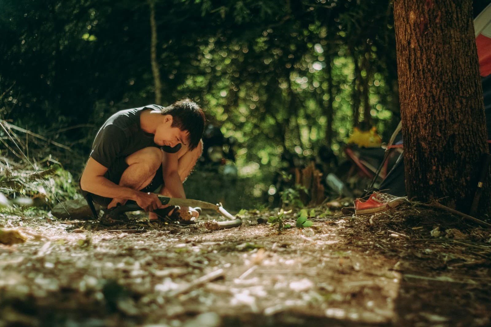 A man crouched on the forest floor cutting a green branch with a folding saw beside a tent