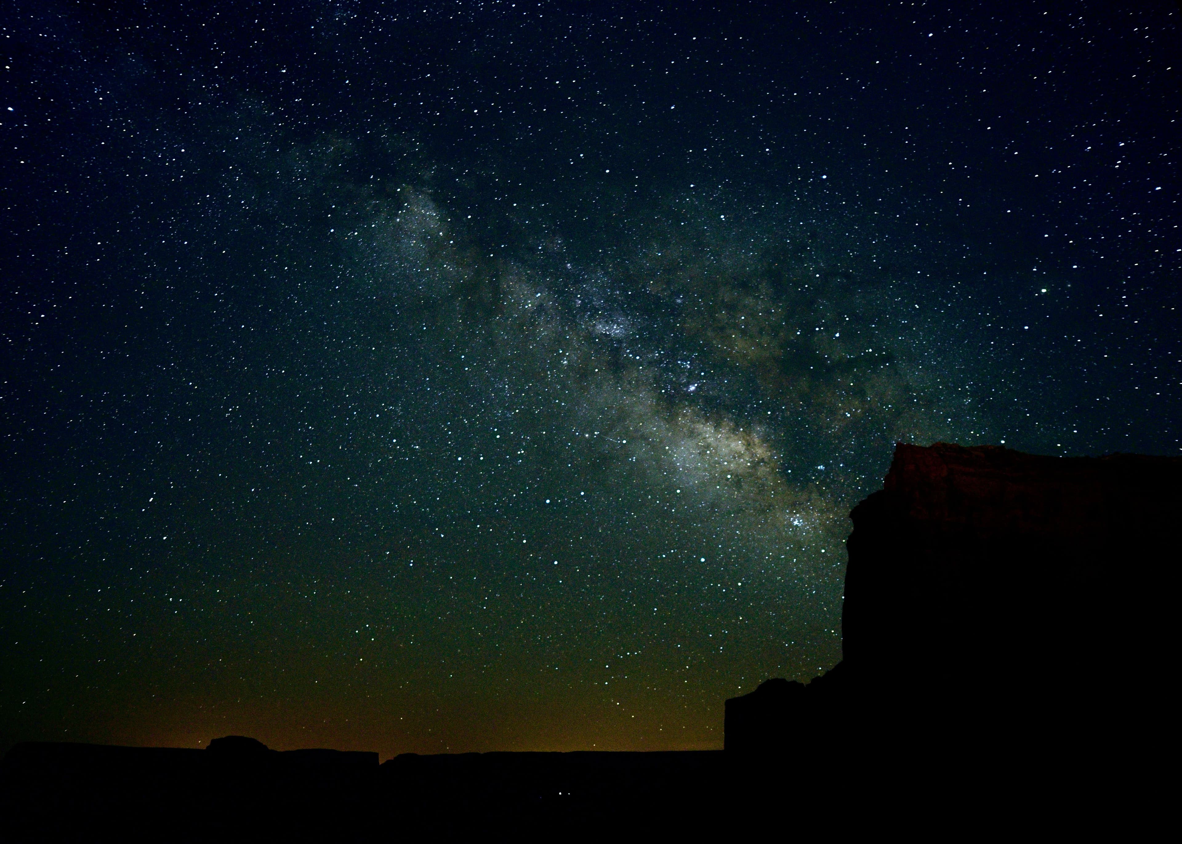 The Milky Way over a silhouetted desert mesa with a faint glow on the horizon