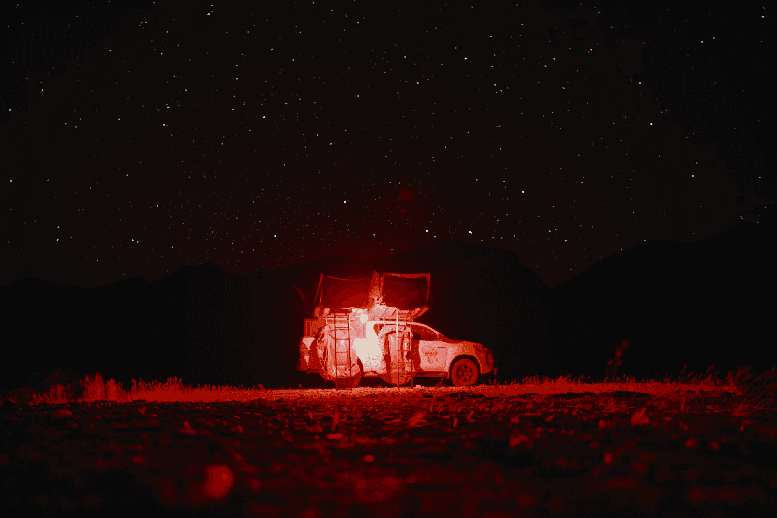 A pickup with rooftop tents lit red below a mountain ridge and starry sky