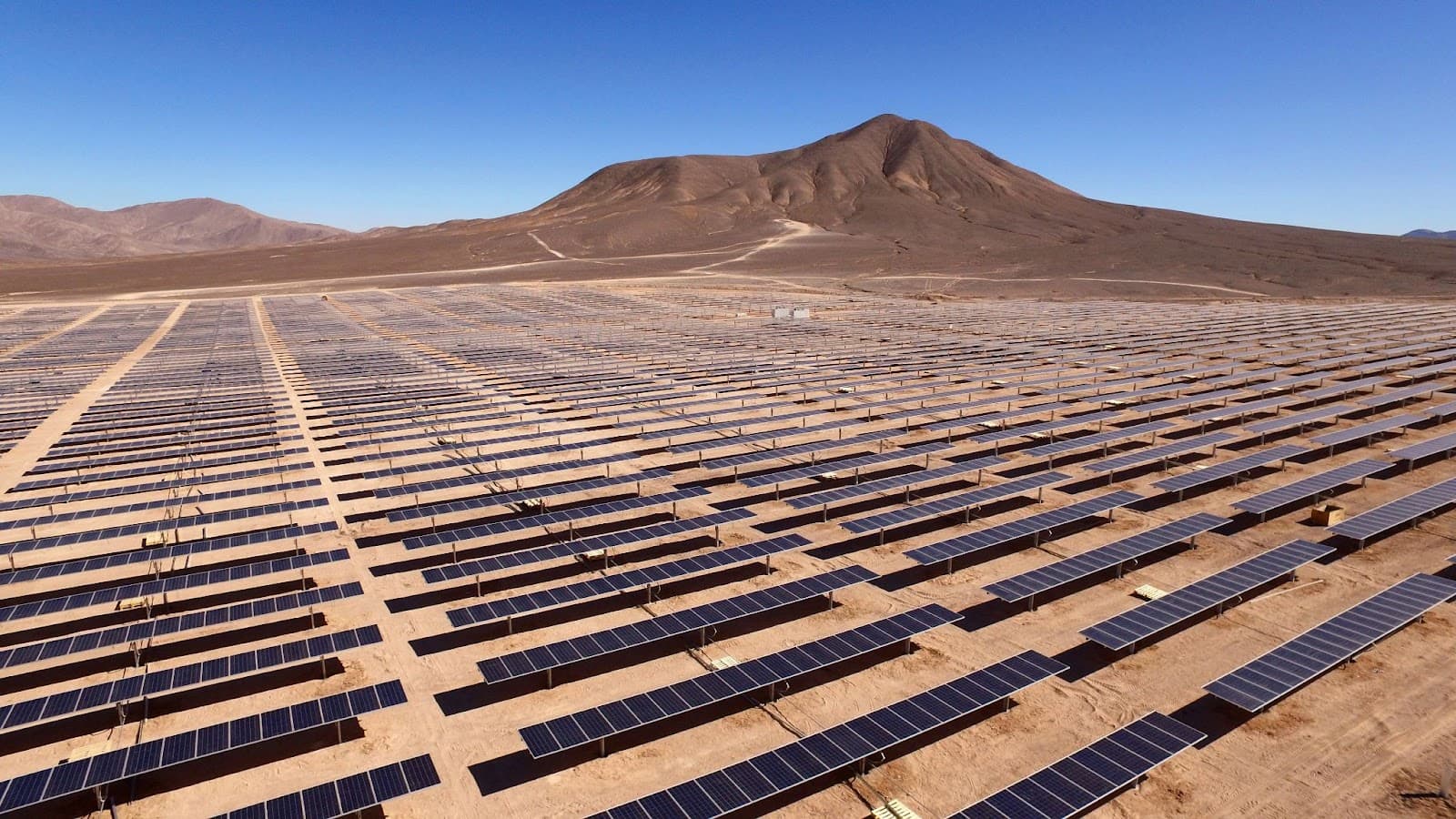 Rows of solar panels covering a desert valley floor beneath a bare brown mountain under a clear sky
