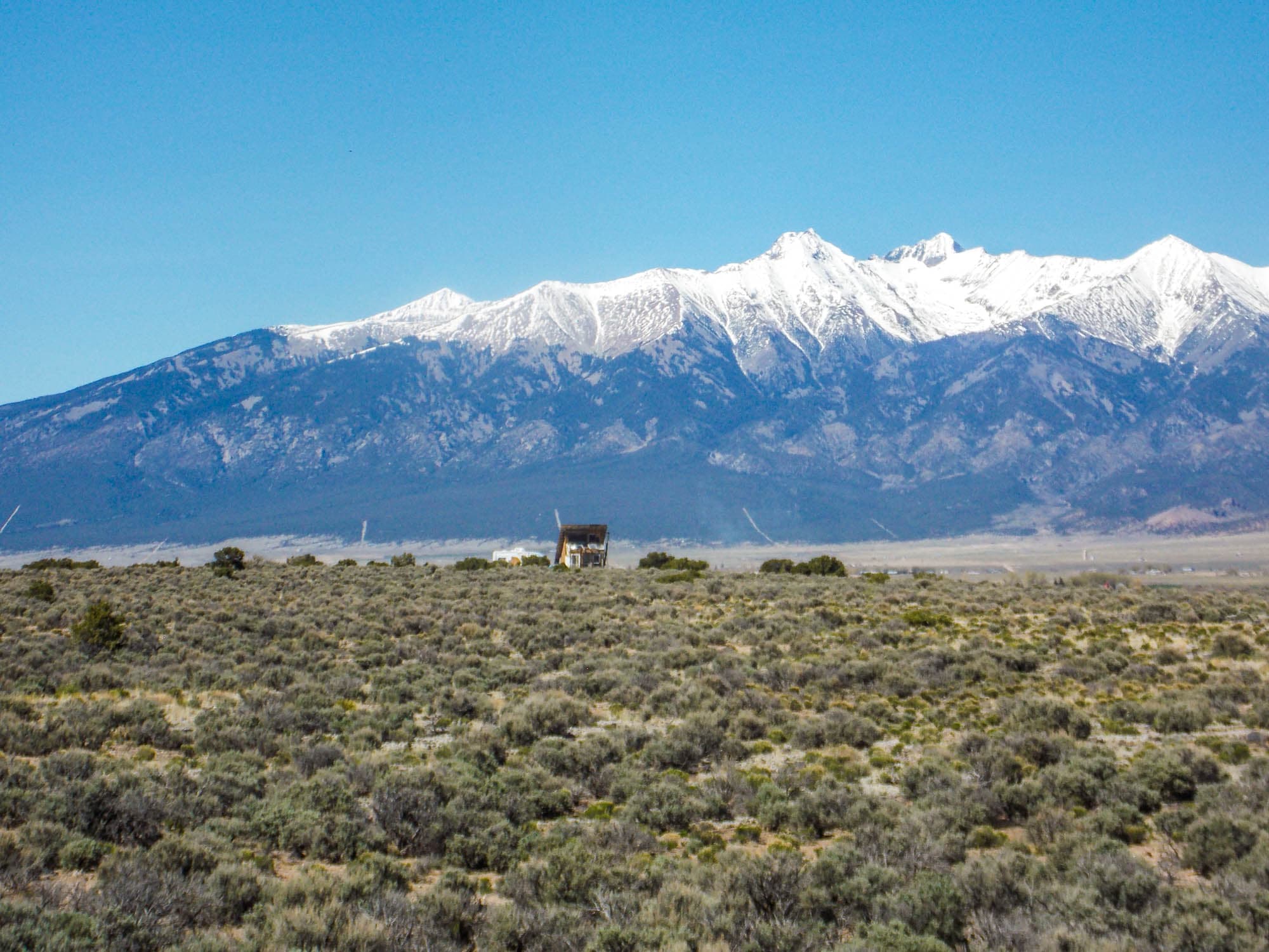 A small cabin standing alone on sagebrush ground in Costilla County, the snow-covered Blanca Peak massif filling the horizon behind it under a clear sky.