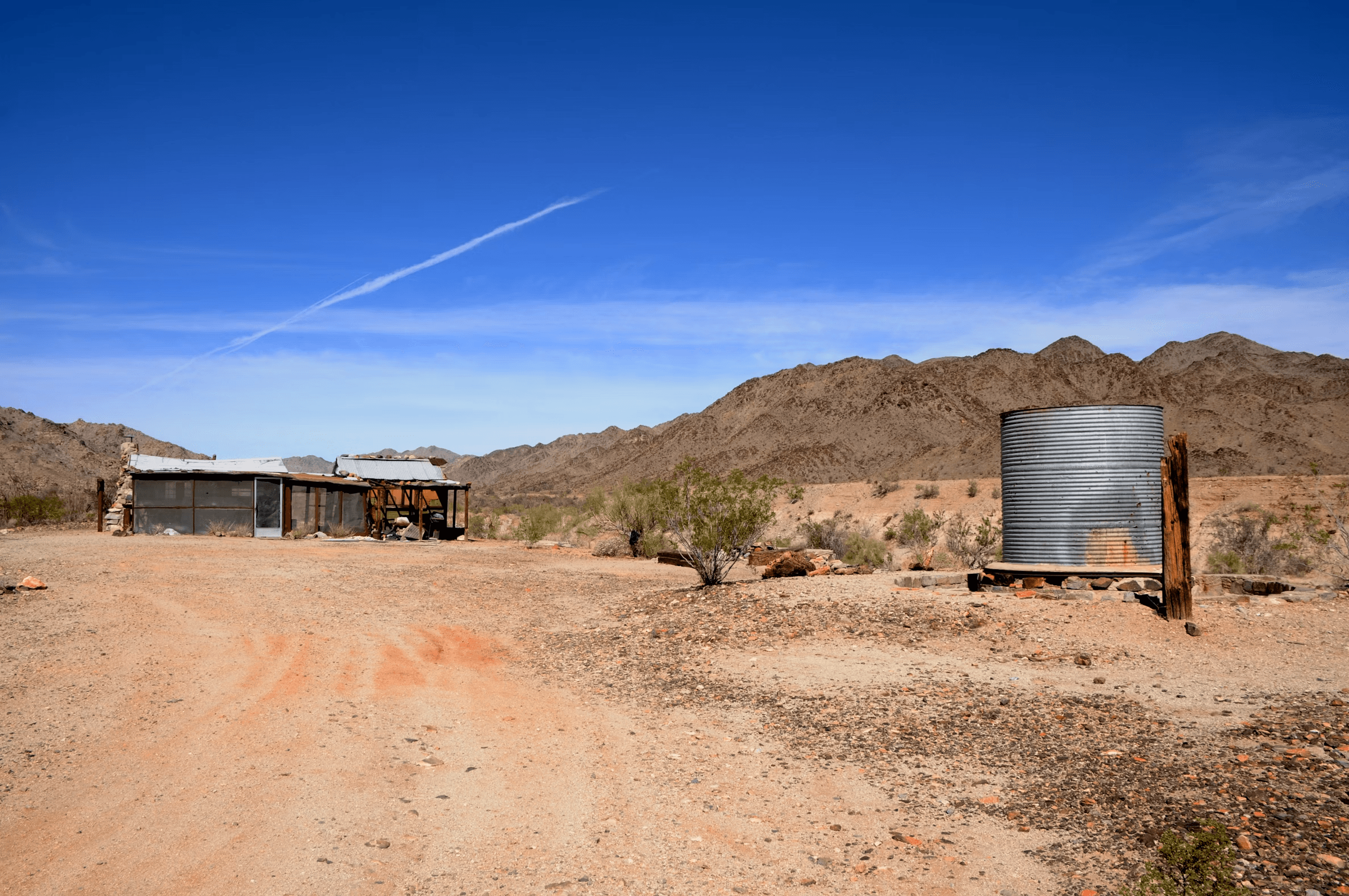 A small desert homestead: a corrugated metal water tank on a timber stand beside an open-sided shelter, with a graded dirt track running past both toward bare rock mountains.