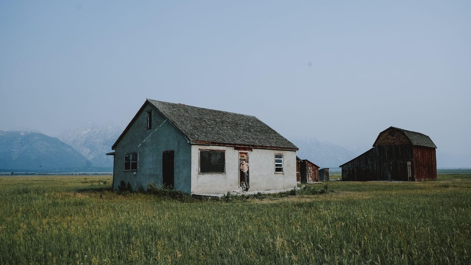 A small white farmhouse and a weathered red barn standing in tall grass with a hazy mountain range behind them
