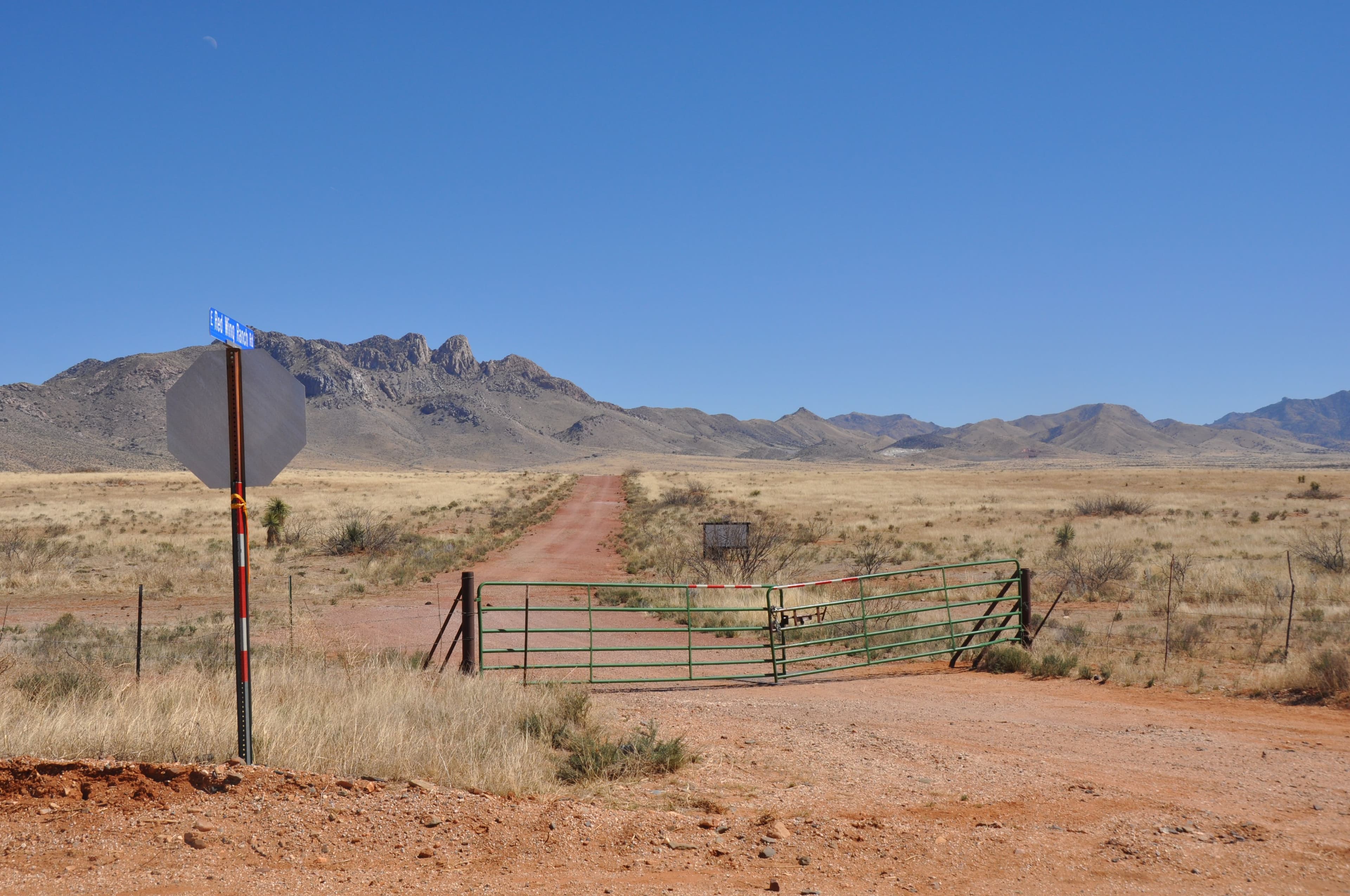 A steel ranch gate standing open on a red dirt track running toward a rugged desert mountain range, a stop sign at the roadside.