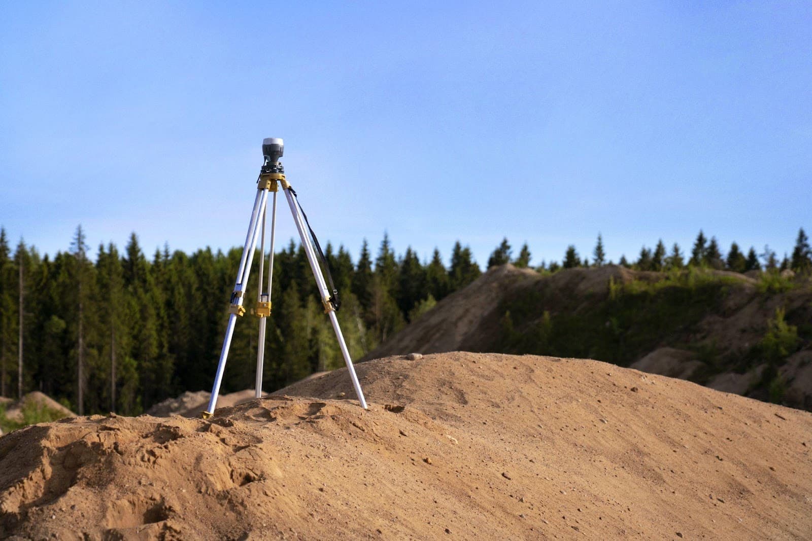 A surveyor total station on a tripod standing on bare graded ground against a blue sky and a distant treeline.