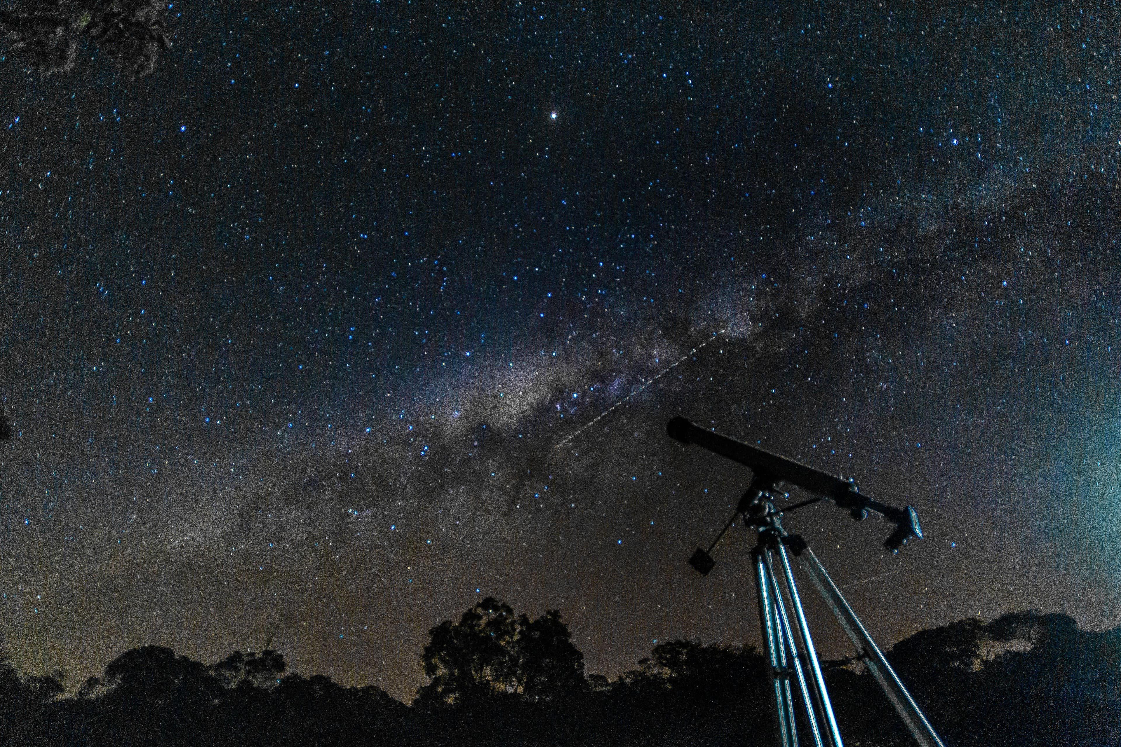 A telescope on a tripod aimed at the Milky Way above a dark treeline