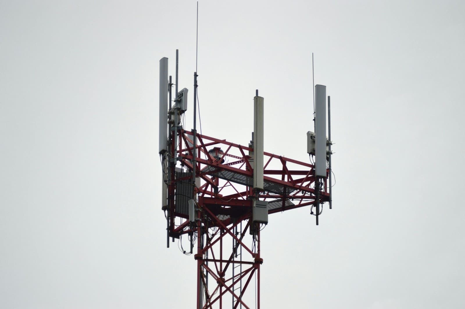 Top of a red and white lattice cell tower, panel antennas mounted on every face, against a flat gray sky