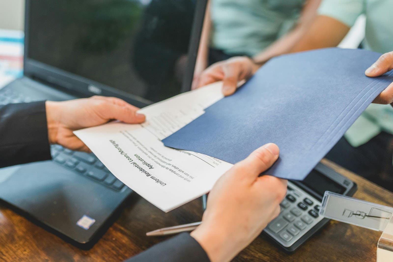 Two people at a desk passing a navy folder of documents across a laptop and a calculator.