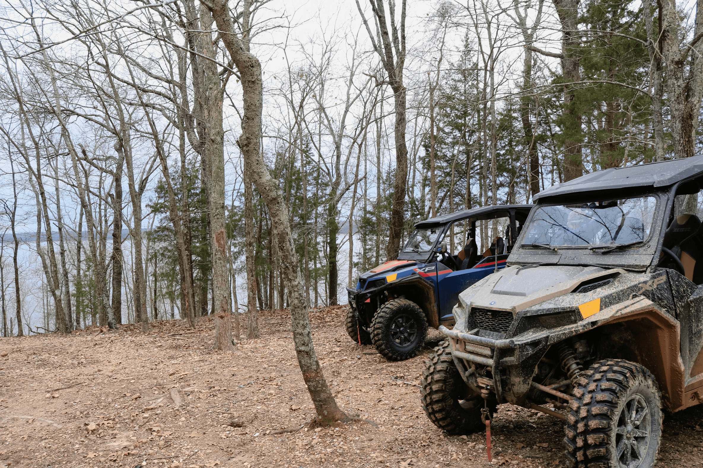 Two side-by-sides parked on a dirt track in a stand of bare hardwoods, one splashed with mud.