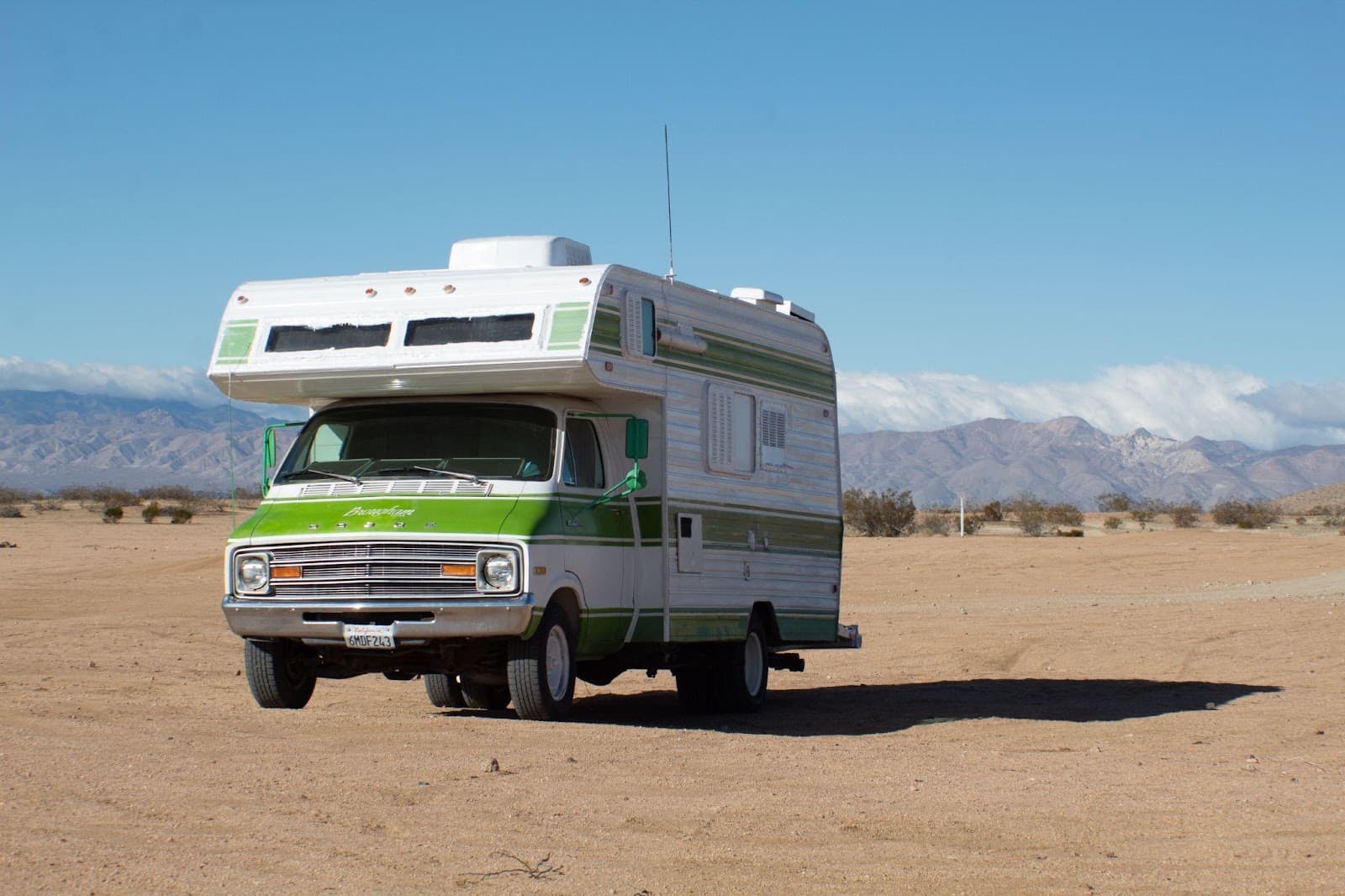 A vintage class C motorhome parked alone on flat open desert ground with a low mountain range behind it.