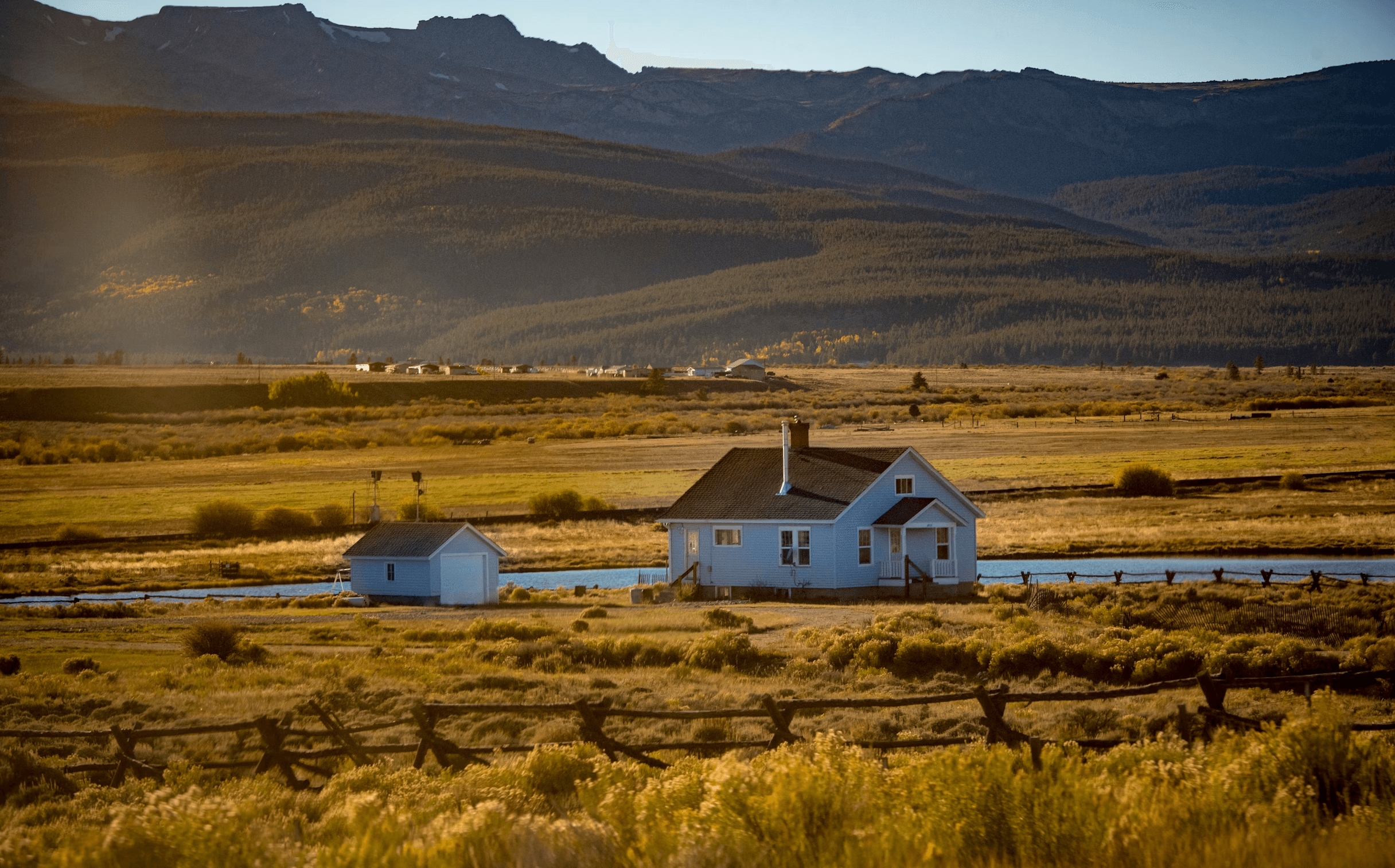 A white farmhouse and outbuildings standing alone in a wide high-desert valley at golden hour, dry grass in the foreground and low hills behind.