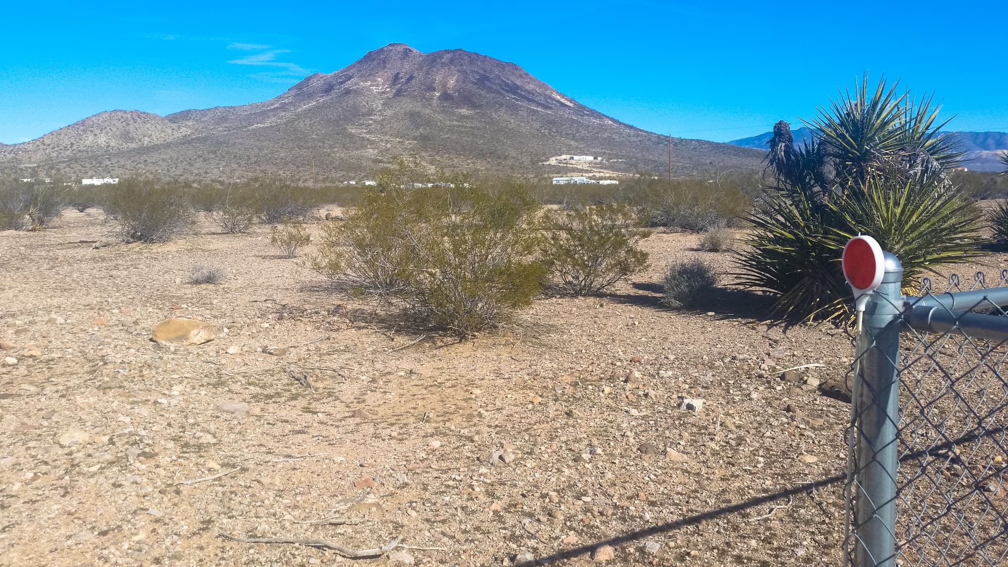 A wire fence corner post with a red cap in open desert scrub, a dark volcanic peak rising behind.